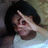 Pixelated image of a person with dark hair making a hand gesture near their face, wearing light-colored clothing against a muted background.