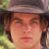 A pixelated close-up of a person's face with light brown hair, wearing a dark cap and visible green foliage in the background.