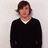 Pixelated static image of a person with brown hair wearing a black sweater over a white collared shirt against a light gray background.