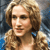 Pixelated portrait of a person with light brown curly hair, wearing a blue earring and light-colored clothing against a blurred background.