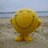 A pixelated yellow figure with a wide smile stands on a sandy beach under a light blue sky.