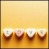 Four heart-shaped candies with letters L, O, V, E arranged horizontally on a yellow background.