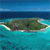 Pixelated static image of a small island with green terrain and a dark cave-like opening, surrounded by turquoise water under a blue sky.