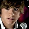 A close-up of a person with light brown hair wearing a white collared shirt and dark jacket, holding a silver microphone against a dark background with purple accents.