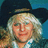 Pixelated static image of a person with long light-colored hair wearing a dark hat and white shirt against blue background.