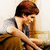 A person with brown hair in a gray shirt and brown pants sits on a couch, resting their head on one hand while looking down.