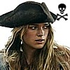 A person with long wavy blonde hair wears a black tricorn hat and a brown jacket, with a black skull and crossbones symbol in the top-right corner.