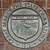 Circular metallic emblem with concentric rings and a central design on a brown textured background.