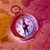 A pixelated circular icon with a central light blue and white pattern surrounded by a dark red ring, set against a gradient background of orange and pink hues.