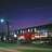 Pixelated nighttime scene with dark blue sky, green ground, and blurred lights from buildings and street lamps.