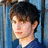 Pixelated portrait of a person with dark hair and light skin wearing a dark blue top against a blue and gray background.