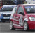 A pixelated static image shows a red vehicle with visible doors and windows, positioned on a gray surface with other vehicles in the background.