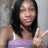 A person with long dark hair wearing a purple tank top makes a peace sign with their right hand against a light gray background.