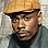 Pixelated portrait of a person wearing a light brown flat cap with dark brim, gray shirt, and black collar visible against blurred background.