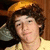 A person with curly brown hair wears a yellow shirt and gray headphones, with a pink heart symbol on the right side of the frame.
