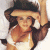 A pixelated image of a woman with long dark hair wearing a white top and wide-brimmed hat, holding her hand near her face against a light background.