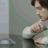 A person in a light-colored long-sleeve shirt leans forward with a clenched fist, positioned near a reflective surface with a rounded metallic object.
