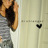 A person with long dark hair wearing a black-and-white striped sleeveless top and light blue jeans stands near a white door, hand raised to face.