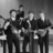 Black-and-white pixelated image of four figures in dark clothing standing in a row, with one figure holding a microphone.