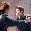 Four show characters in a tense interaction with one character holding another's shoulders in a dimly lit indoor setting