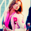 A person with long light brown hair wears a white jacket with pink accents and a dark top, standing against a blurred background with light and dark tones.