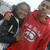 Two people pose together, one in a red hoodie with white text, the other in a black jacket with white stripes, against a light background.