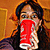 A person with long dark hair holds a red cylindrical object in front of their face, set against an orange-brown background.