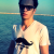 Pixelated image of a person wearing dark cap and white shirt with black mustache graphic, standing against blue water background.