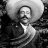 Black-and-white pixelated image of a person with a mustache wearing a dark jacket and light shirt, set against a blurred background with a large circular shape.