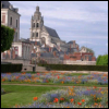 A static image of a historic building with a tall tower, surrounded by a flower garden with orange and blue blooms under a partly cloudy sky.