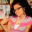 A pixelated image of a person with dark hair wearing a pink and white patterned top, holding a rose, with a blurred background containing indistinct shapes and colors.