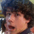 Pixelated close-up of a person with dark curly hair and light skin, mouth open in mid-expression against a blurred background.