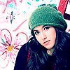 A person with long dark hair wears a green knit beanie and dark clothing against a colorful floral background with pink and blue patterns.