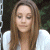Pixelated image of a person with long brown hair wearing a white top against a dark background with green foliage.