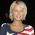 Pixelated image of a person with short blonde hair wearing a dark blue top with white and red stripes on the shoulder against a dark background.