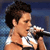 Pixelated image of a person with dark hair holding a microphone, wearing a white sleeveless top against a blue background.