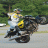 A pixelated static image shows a person riding a motorcycle on a paved road with green foliage in the background.