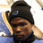 A person wearing a dark beanie with a blue circular symbol, light-colored earflaps, and a dark turtleneck against a blue and white background.