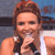 A person with light brown hair holds a black microphone close to their mouth, wearing a white sleeveless top against a blurred background.