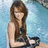 A woman with long brown hair sits holding a black steering wheel against a blue water background.