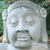 Pixelated grayscale image of a stone or marble face with closed eyes, a broad nose, and a mouth showing teeth, set against a blurred green and brown background.