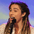 A person with long brown hair and a white top sings into a black microphone against a blue gradient background.