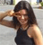 A person with long dark hair wearing a sleeveless dark top, raising their right arm to their forehead in a saluting gesture against a blurred background.