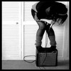 Black-and-white photo of a person in dark clothing bending over a box, with legs spread and hands near the face, against a louvered door background.