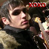 A person with short dark hair and dark eye makeup wears a black jacket with a fur-lined collar, holding a guitar with a checkered pattern, while red 'XOXO' text appears in the top-right corner.
