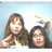 Two people pose against a light blue background with one person making a peace sign and the other raising their hand in a wave gesture.
