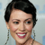 A pixelated close-up of a person with short dark hair, light skin, and a broad smile showing teeth, wearing dangling earrings against a light blue background.