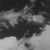 Grayscale abstract composition with dark and light pixel clusters forming indistinct shapes against a gradient background.
