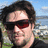 Pixelated image of a person with dark hair and sunglasses against a blue sky background with blurred green and gray elements.
