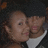 Two people in close proximity, one with light skin and short hair wearing a white shirt under dark clothing against a dark background.